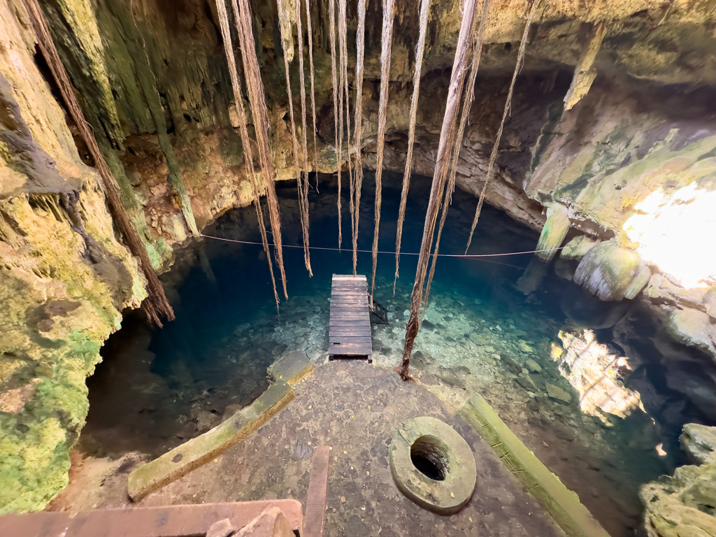 Die grosse Celote von oben gesehen. Sonnenlicht und Wurzelwerk ranken sich hinab. Ein kleiner Brunnen und der Steg ins blaue klare Wasser.