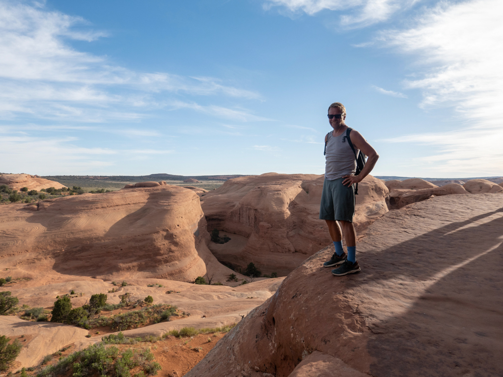 Jo posiert am Wegesrand zum Delicate Arch. Unten eine felsige Ebene mit grünen Flecken dazwischen