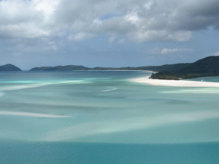 wie sanfte Wellen zeichnet sich der weisse Sand unter dem flachen Wasser ab. Weiter leerer Strand mit ein paar wenigen Booten in der Bucht