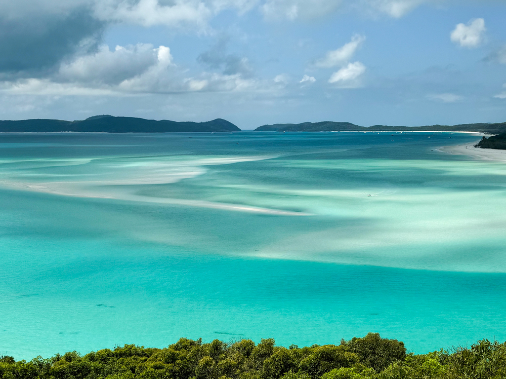 Südseepanorama:wie sanfte Wellen zeichnet sich der weisse Sand unter dem flachen Wasser ab. Weiter leerer Strand mit ein paar wenigen Booten in der Bucht