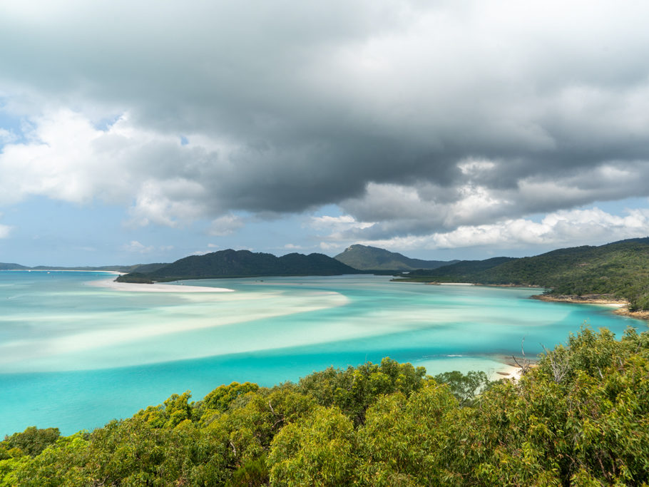 Südseepanorama:wie sanfte Wellen zeichnet sich der weisse Sand unter dem flachen Wasser ab. Weiter leerer Strand mit ein paar wenigen Booten in der Bucht
