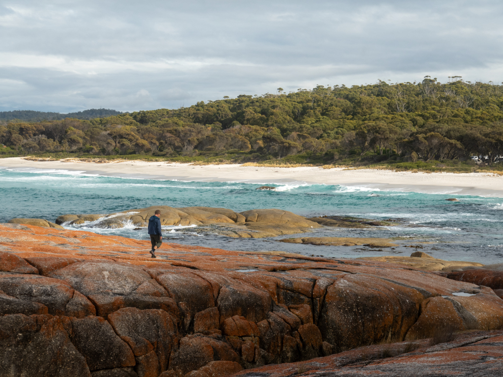 Mit Jo auf den grossen mit roten Flechten bewachsenen Granitfelsen unterwegs. Dahinter der weisse Strand der Bay of Fire