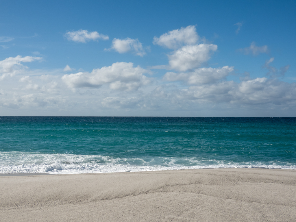 Wie ein gemaltes Bild. Vorne der weisse Stand mit dem blauen Meer und ein paar Schäfchenwolken am Himmel.